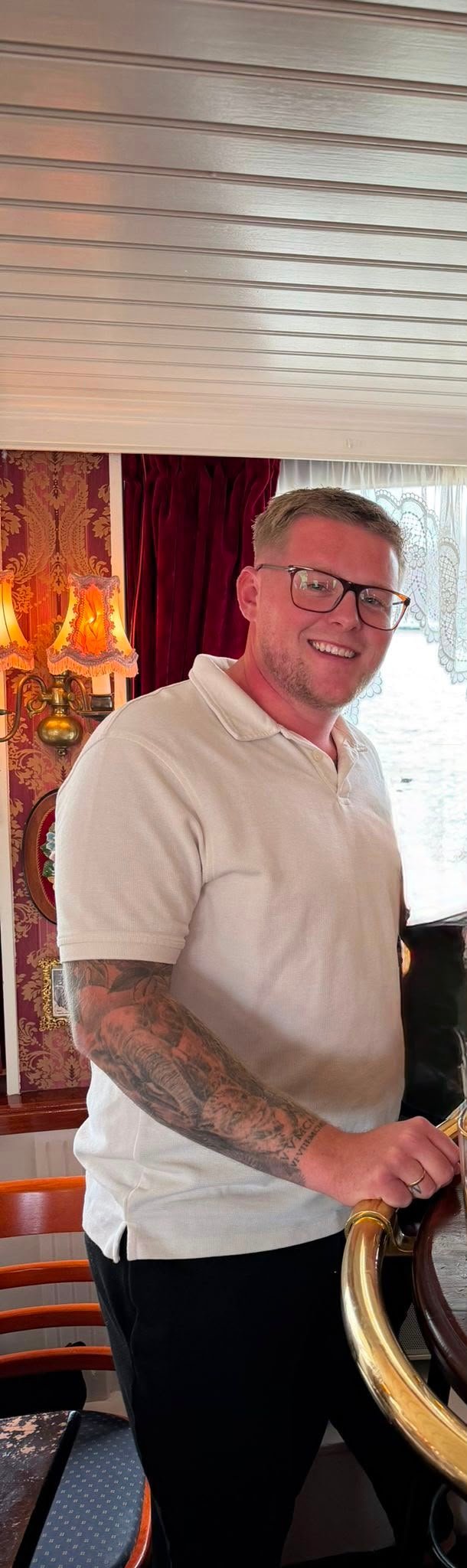 Man with glasses and arm tattoo smiling indoors in white polo shirt and dark pants